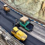 Vibroacabadora e rolos compactadores aplicando asfalto em trecho de rodovia, vistos do alto