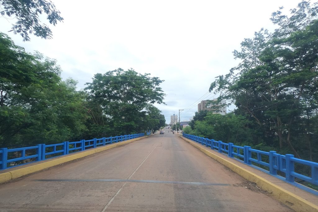Ponte com guarda-corpo azul sobre córrego arborizado em Rondonópolis