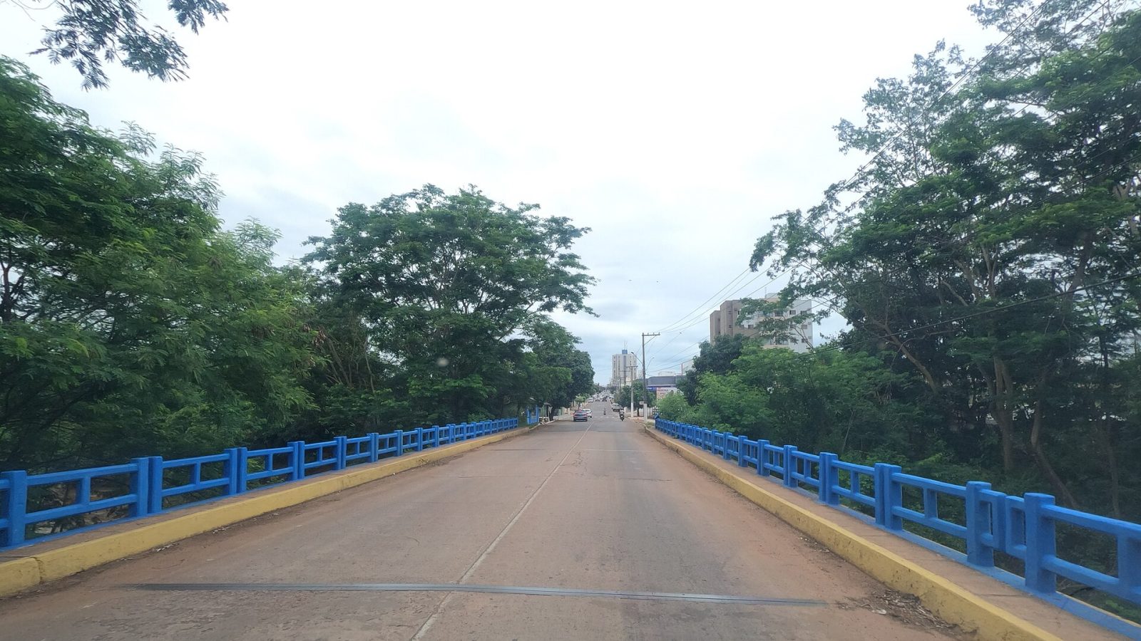 Ponte com guarda-corpo azul sobre córrego arborizado em Rondonópolis