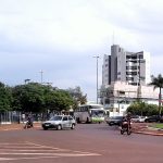 Rotatória em Tangará da Serra, com ônibus, carros, motos e prédios ao fundo