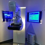 Equipamento de mamografia em sala de exame, com monitores iluminados ao fundo