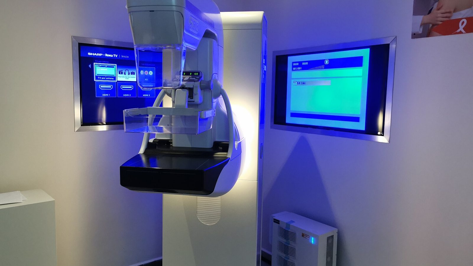 Equipamento de mamografia em sala de exame, com monitores iluminados ao fundo