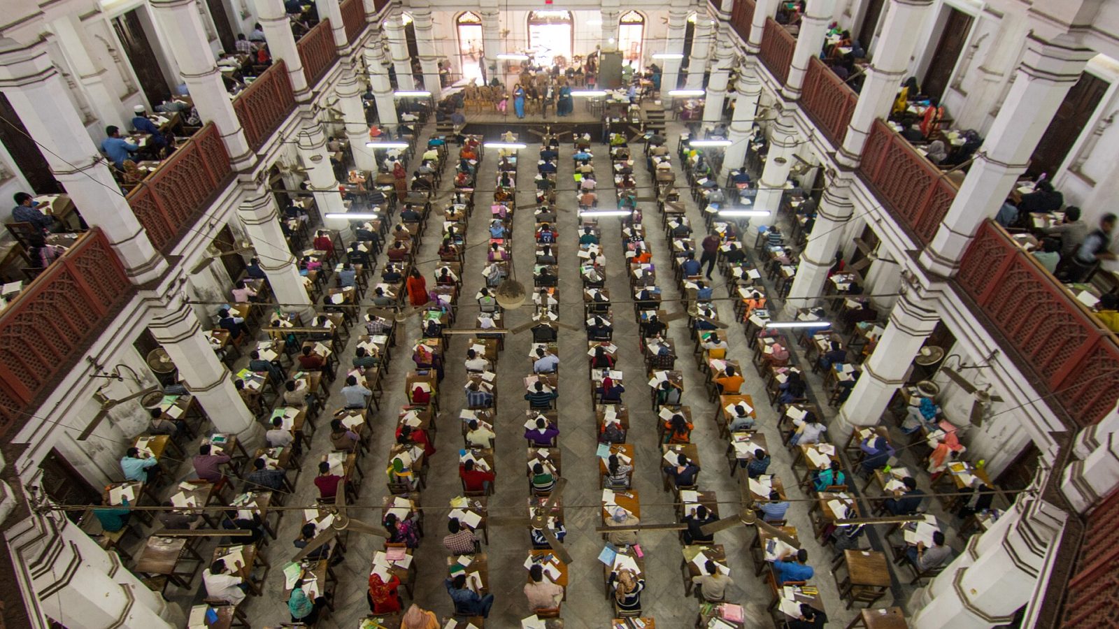 Vista do alto de um grande salão com centenas de candidatos sentados em fileiras de carteiras durante uma prova