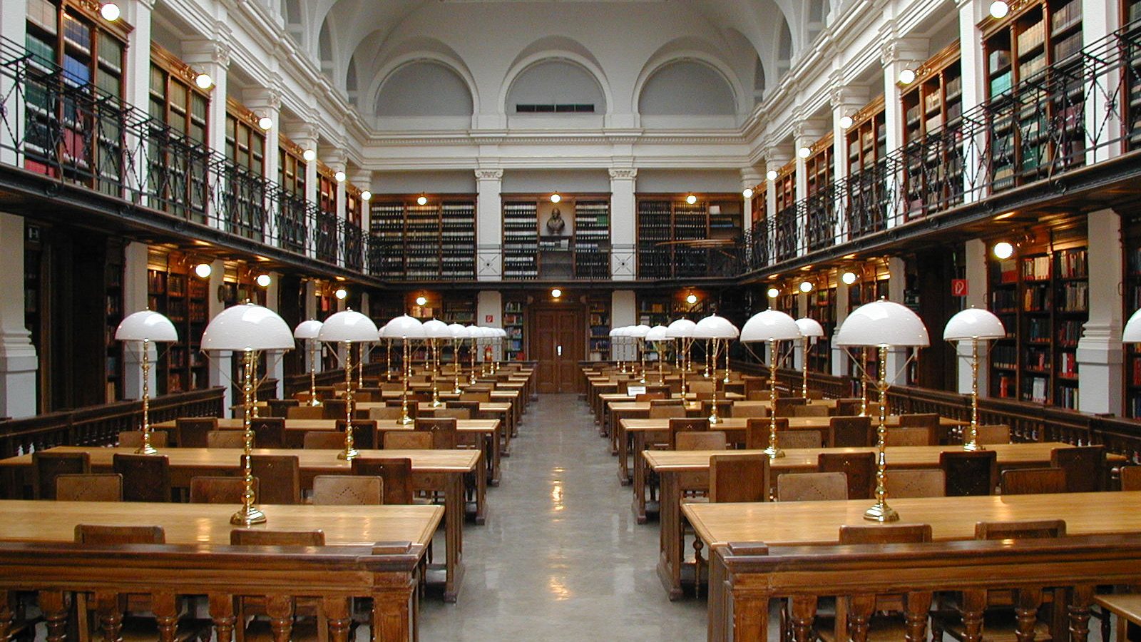 Sala de leitura de biblioteca universitária com mesas, luminárias e estantes de livros