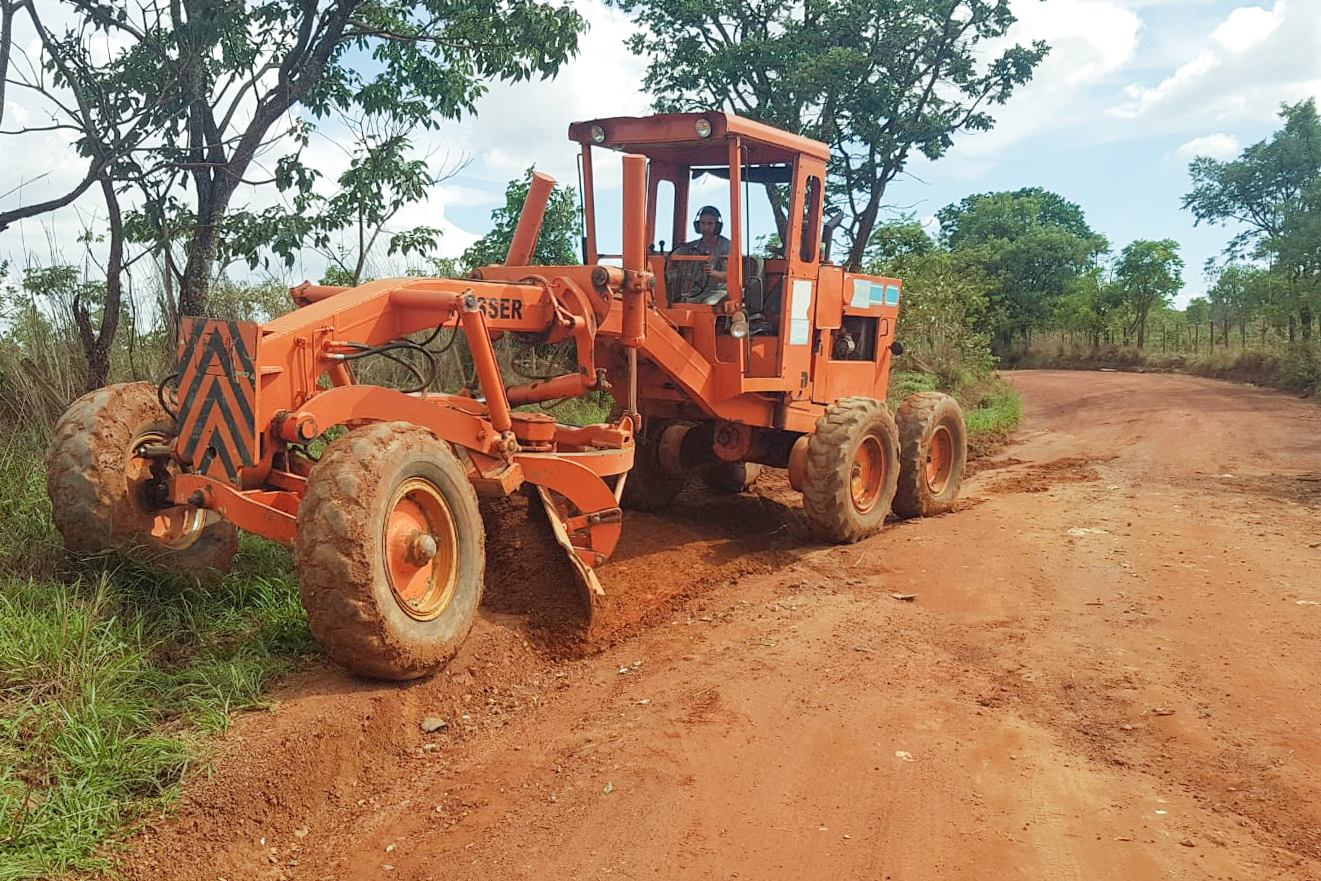Motoniveladora alaranjada raspa e nivela estrada de terra vermelha em área rural