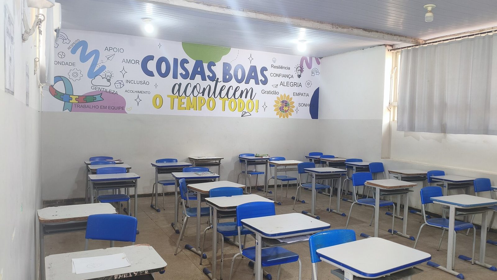 Sala de aula de escola pública com carteiras azuis vazias e painel colorido com a frase Coisas boas acontecem o tempo todo