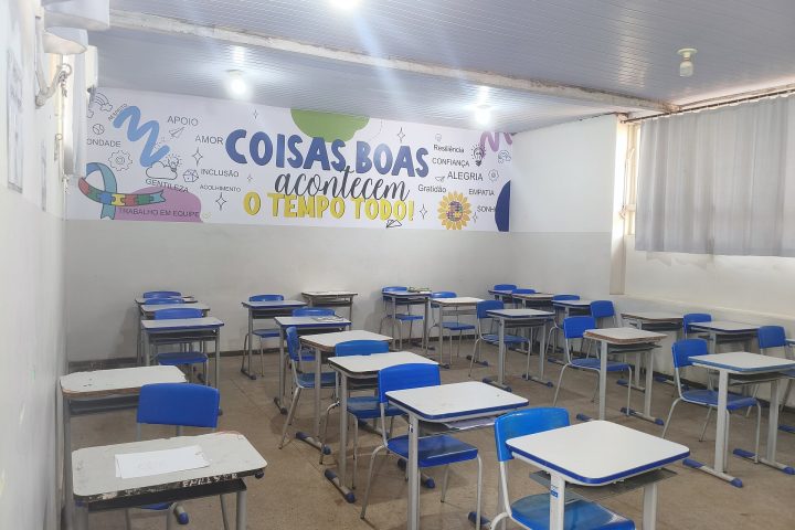 Sala de aula de escola pública com carteiras azuis vazias e painel colorido com a frase Coisas boas acontecem o tempo todo