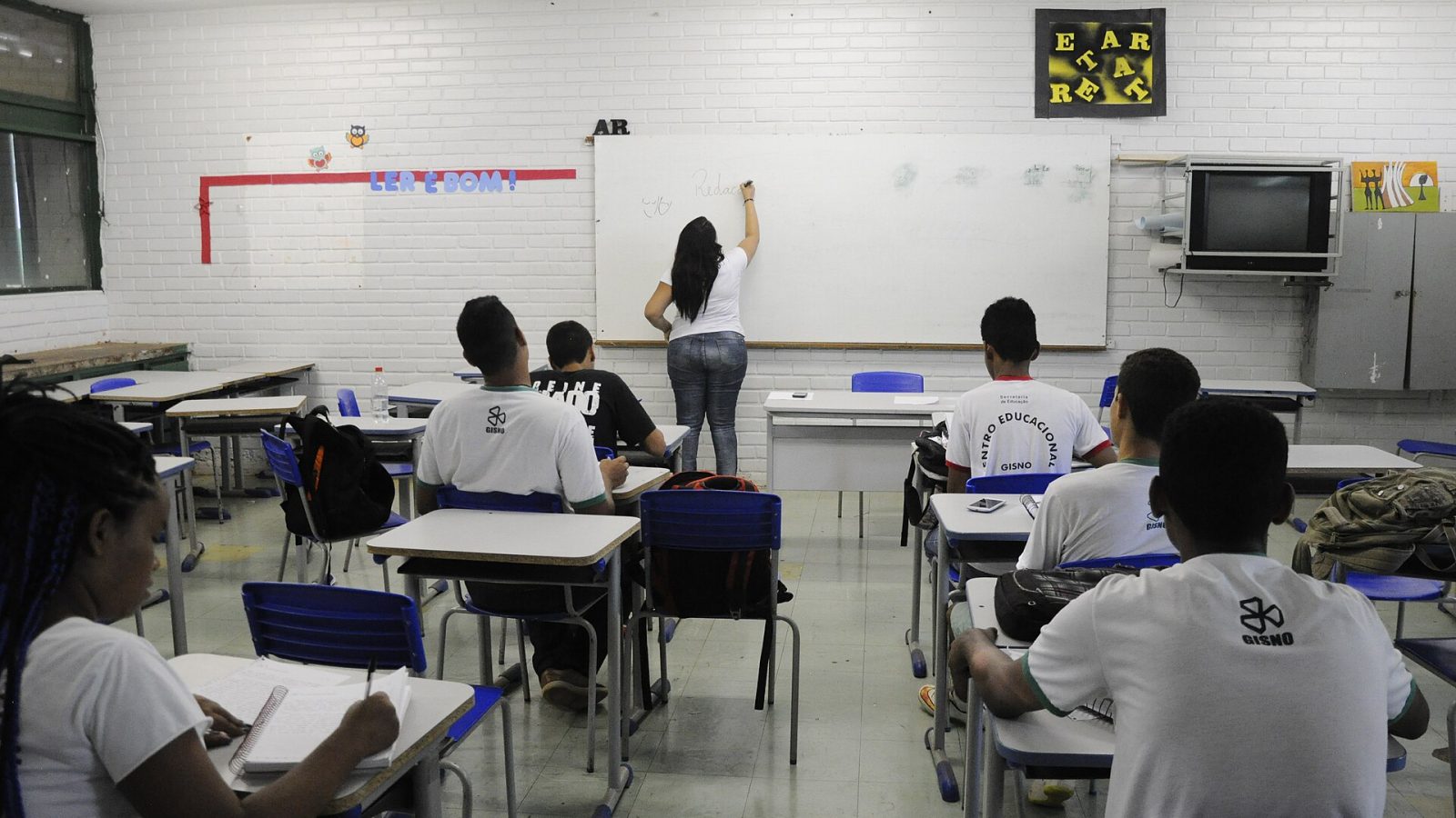 Professora escreve no quadro branco enquanto estudantes acompanham a aula em sala de escola pública