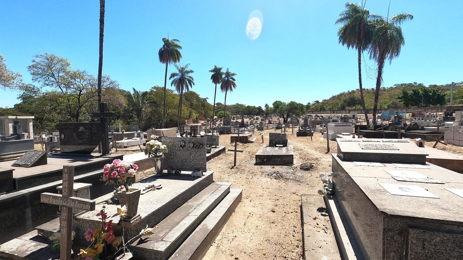 Túmulos e jazigos enfileirados em cemitério sob céu azul, com flores sobre algumas sepulturas