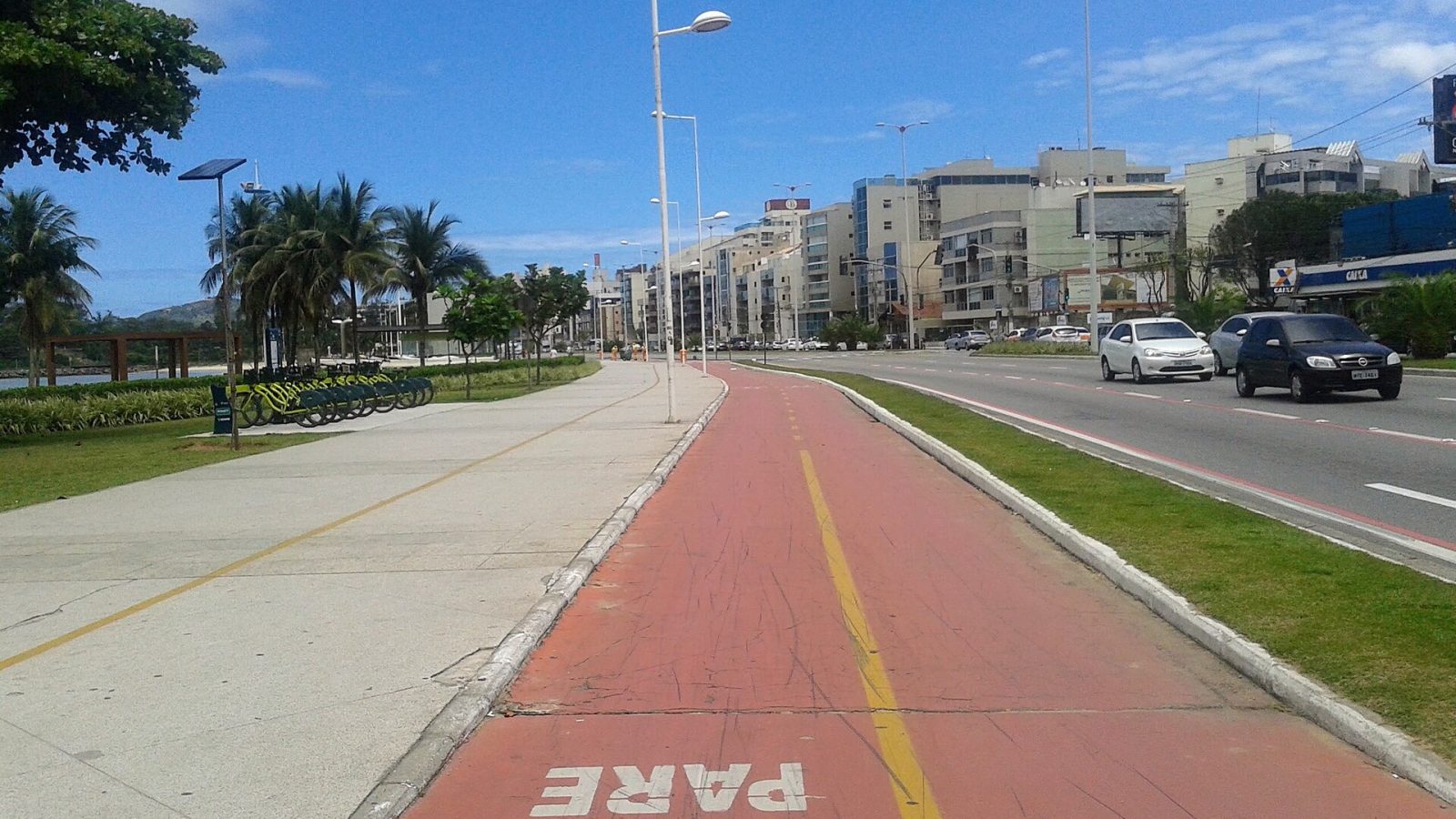 Ciclovia vermelha com a sinalização Pare no piso, entre a calçada e a pista de uma avenida urbana