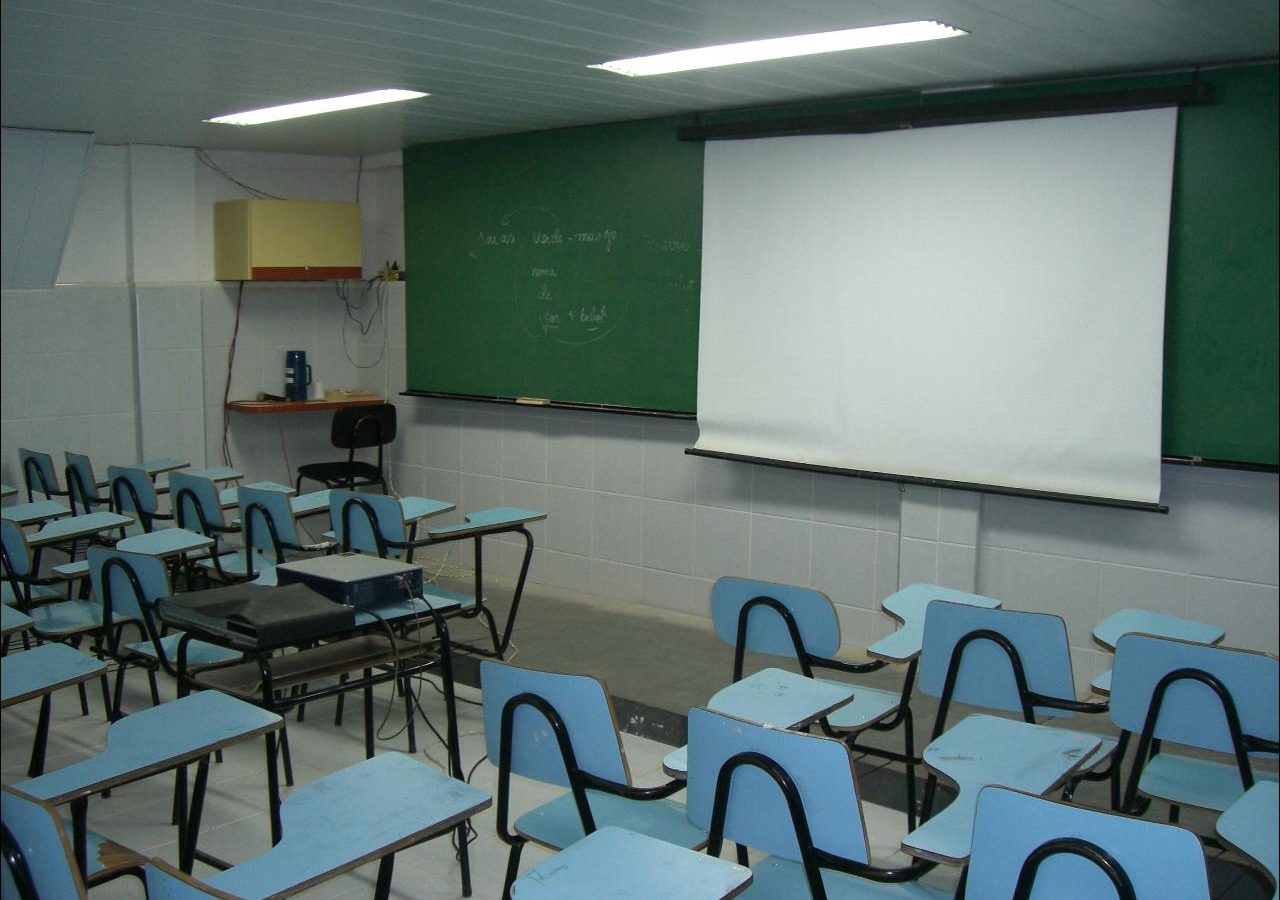 Sala de aula vazia com carteiras azuis enfileiradas, quadro e tela de projeção