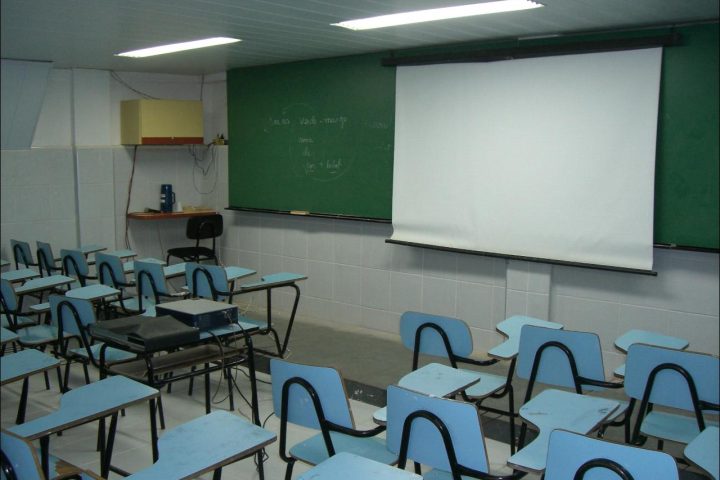 Sala de aula vazia com carteiras azuis enfileiradas, quadro e tela de projeção